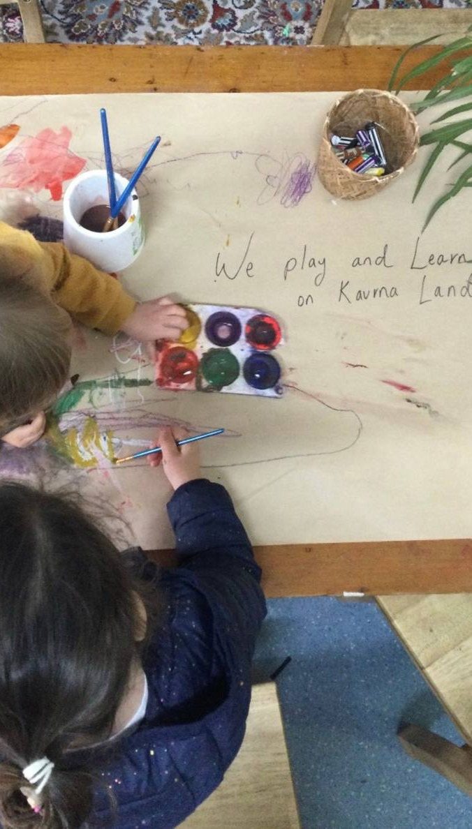 Children painting at a table beneath a handwritten sign reading We play and learn