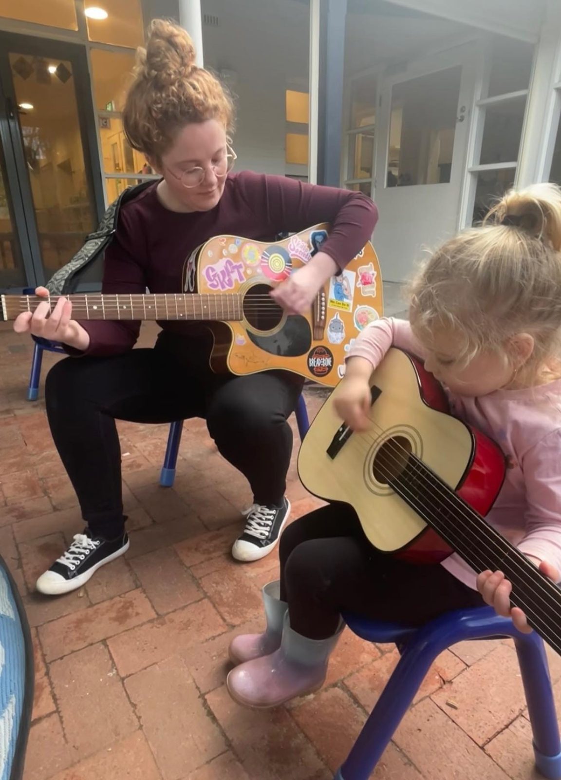 An educator playing guitar with a young child holding a small guitar
